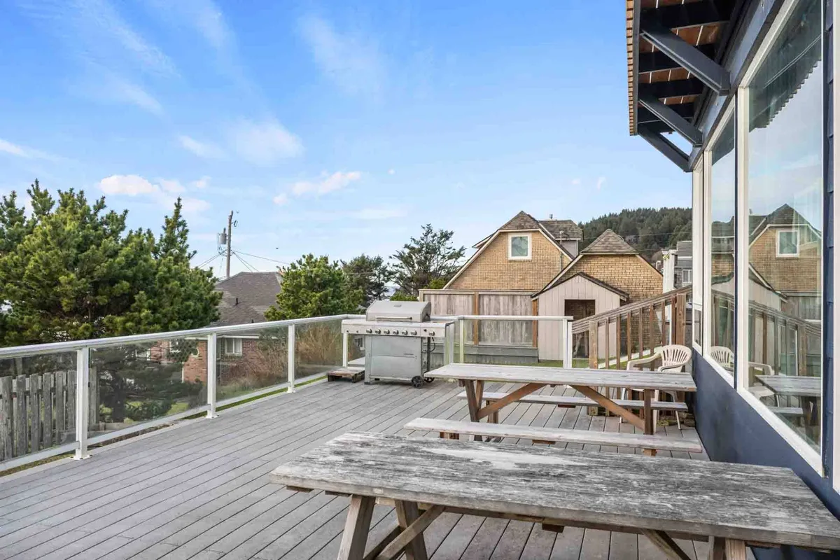An exterior view of a spacious deck with picnic tables, a grill, surrounding trees, and nearby houses. The deck offers plenty of space for outdoor dining, barbecues, and group gatherings, all set within a casual and relaxed atmosphere.