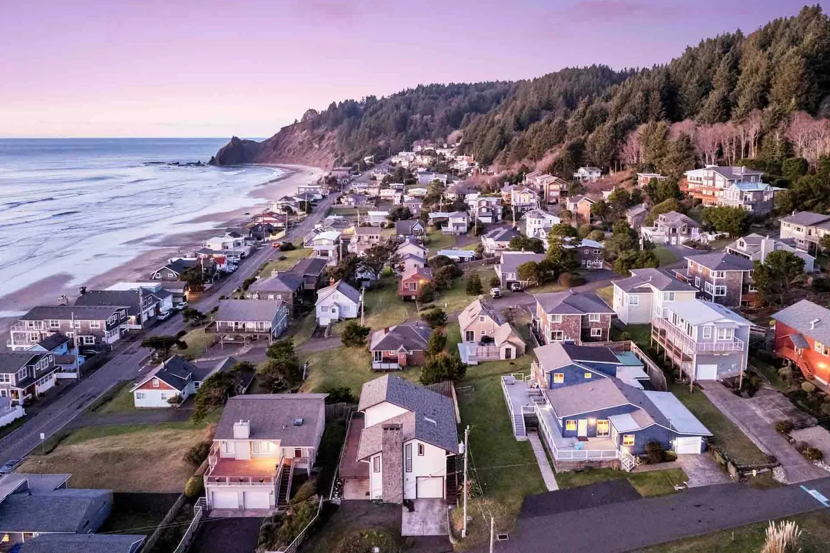 A scenic view of beachfront properties with waves rolling onto the sandy shore and forested hills in the distance. The image captures the inviting coastline just steps from the house, emphasizing the close proximity to the beach and natural beauty of the area.