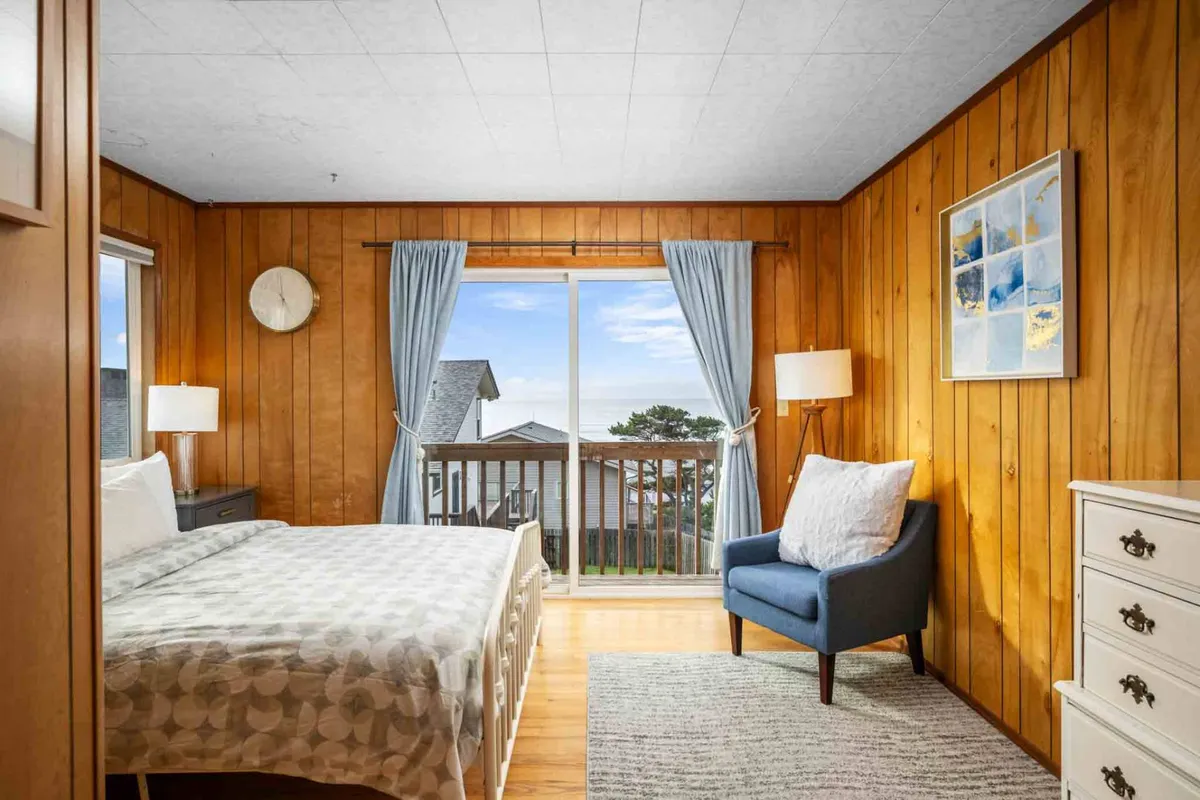 A tranquil bedroom with a bed positioned near a window that looks out towards the ocean, accompanied by a blue accent chair and a white dresser. The soft lighting, calming colors, and cozy setup invite relaxation and peaceful moments.