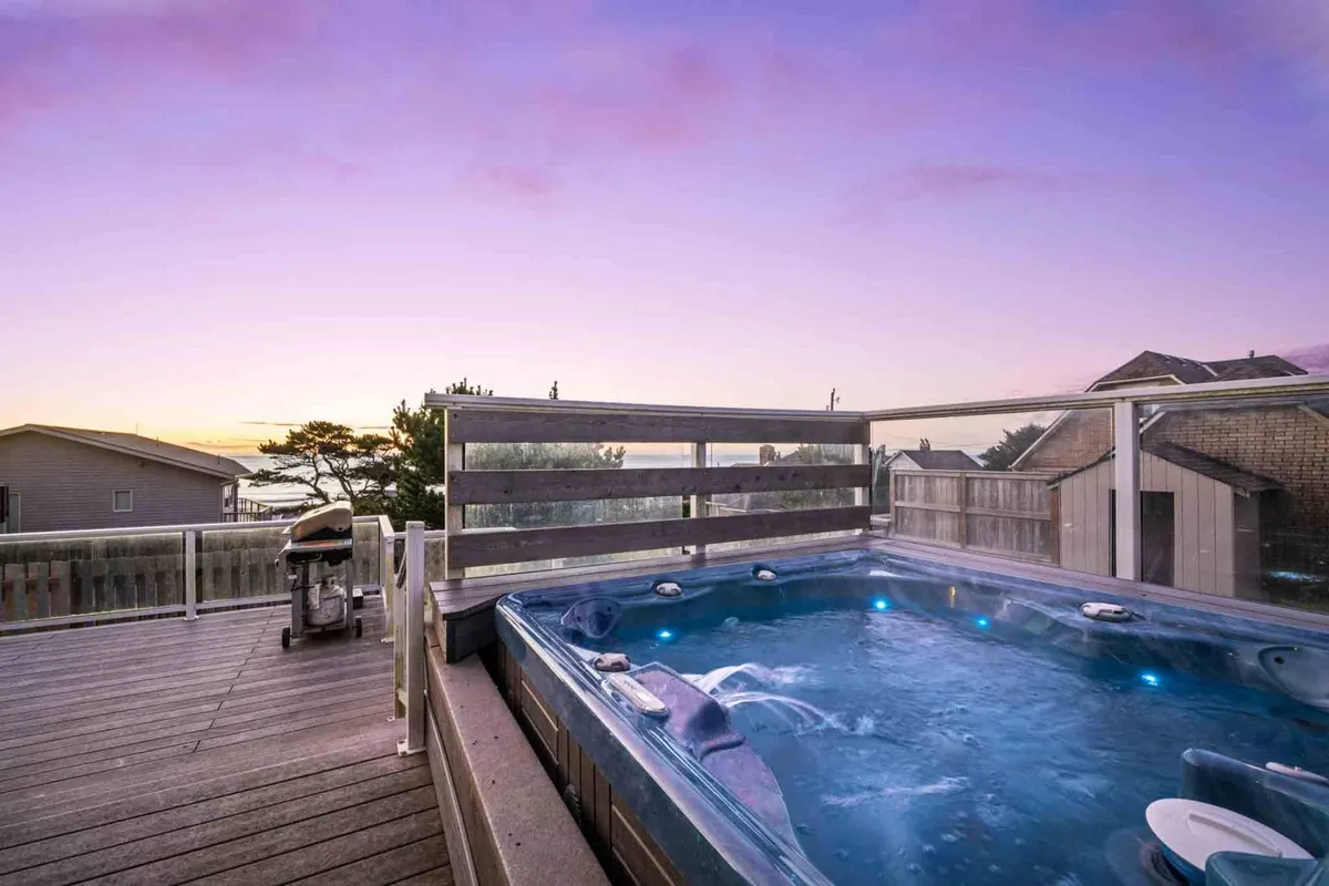 An inviting exterior deck with a steaming hot tub, surrounded by glass wind screens, overlooking the ocean as the sun sets. The deck is spacious, offering room for grilling and relaxing while taking in the view.