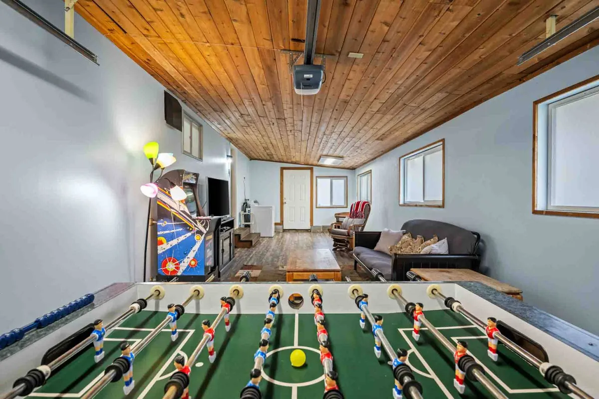 The game room featuring a foosball table, a comfortable sofa, an arcade game, and large windows that bring in natural light. The scene captures guests engaging in foosball and relaxing on the sofa, with the arcade game visible in the background.