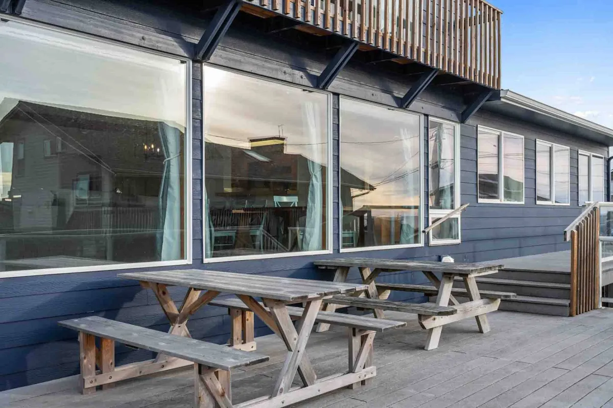 An exterior scene with large windows, wooden picnic tables, and a spacious deck area. The mood is casual and welcoming, suggesting a relaxed, communal outdoor space adjacent to the home.