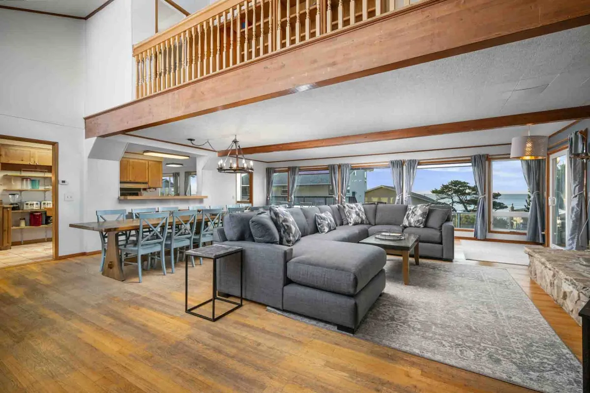 A spacious, sun-filled great room featuring a large sectional sofa and a dining table positioned beside expansive windows. The windows overlook stunning ocean views, and the area exudes an inviting, communal atmosphere. The fireplace adds warmth to the living space, while the furniture arrangement encourages relaxation and togetherness.