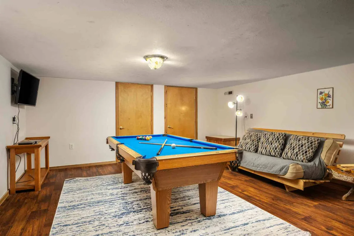 A welcoming game room scene with a pool table at the center, a cozy sofa, modern light fixture, and a television mounted nearby. The ambience is set for game nights or simply relaxing with friends and family.