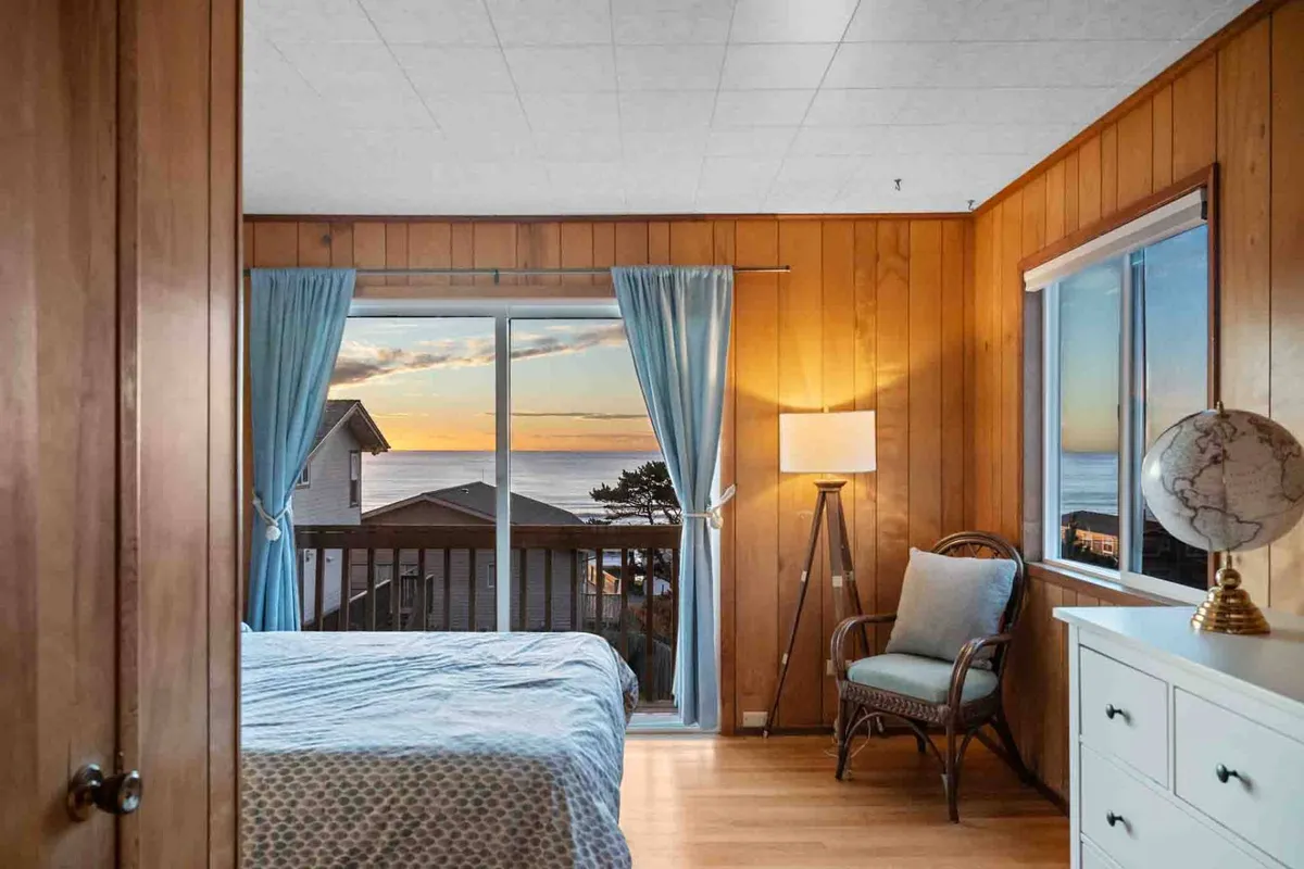 This image captures a serene bedroom with light blue bedding, large windows framing sweeping ocean views, a comfortable chair, and a floor lamp beside a dresser topped with a globe. The tranquil atmosphere is enhanced by natural light, creating an inviting nook for relaxation, reading, or intimate conversation.