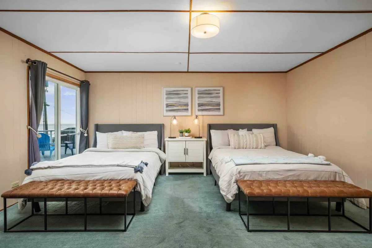 A comfortable bedroom setup featuring two beds, a nightstand with decorative touches, artwork on the wall, and a window that lets in gentle natural light. The twin beds and inviting decor suggest flexibility for families or groups.