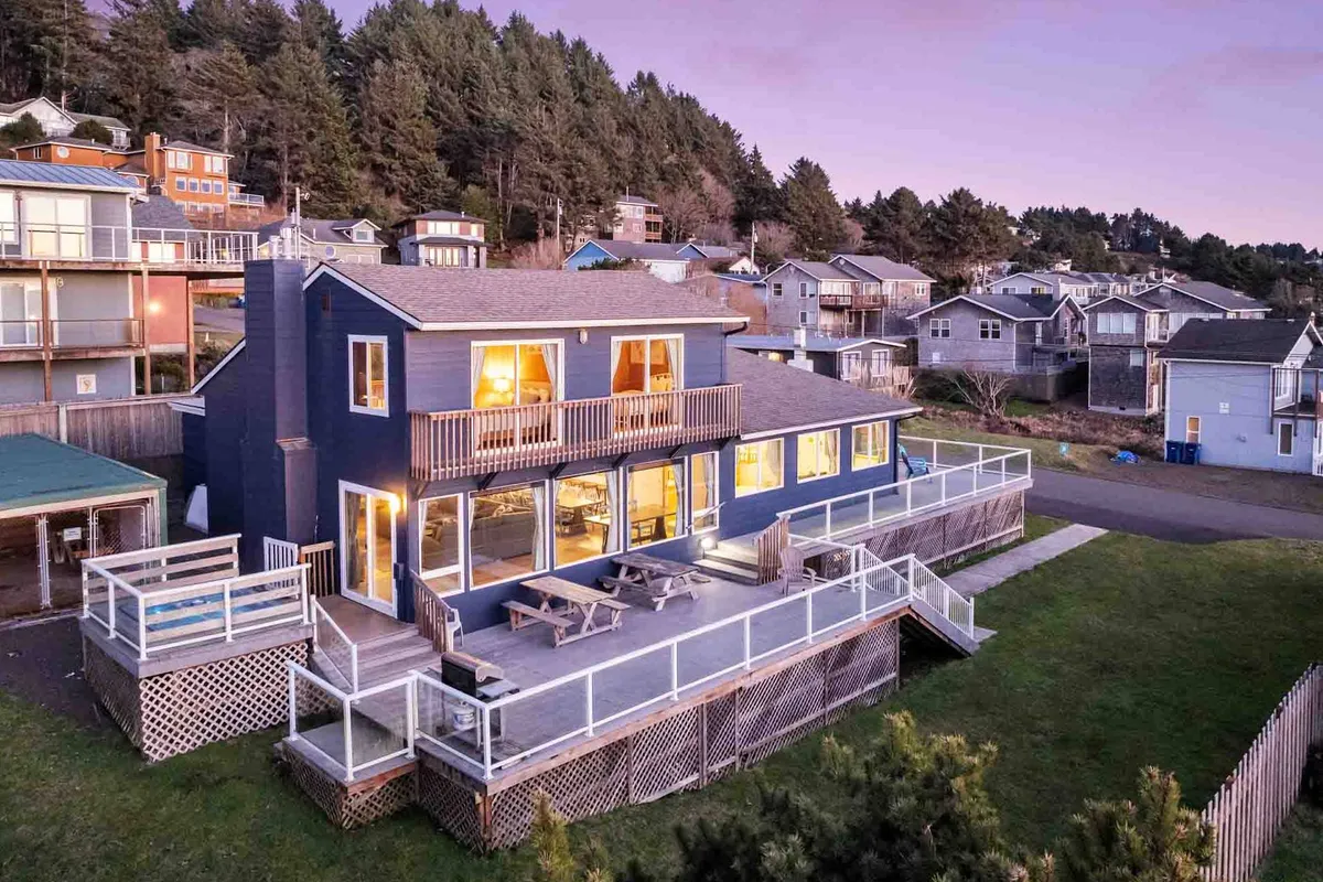 The image features the inviting exterior of a spacious single-family home nestled among lush greenery. Its expansive deck is perfect for gatherings, framed by floor-to-ceiling windows that promise breathtaking views. The scene evokes a tranquil, welcoming atmosphere ideal for families or groups looking to relax together.