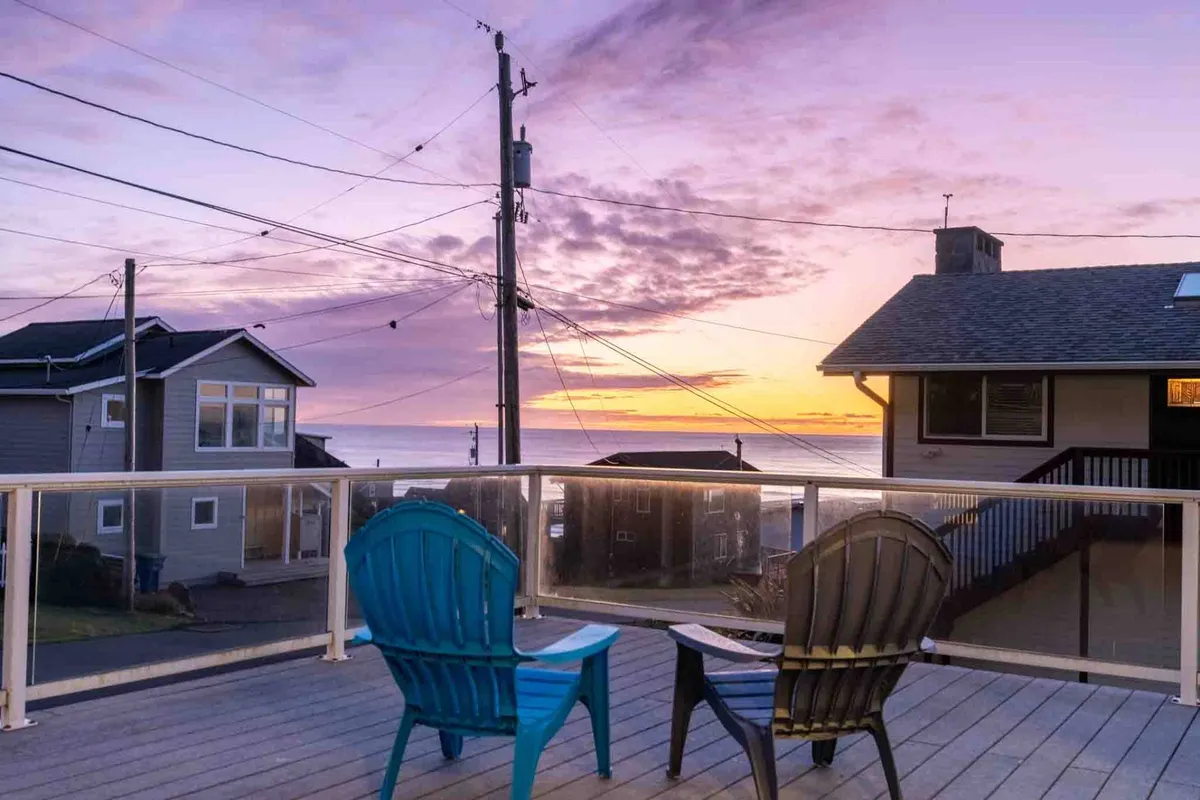 Two Adirondack chairs are positioned on an exterior deck or lawn, facing a dramatic sunset view over the ocean and neighboring houses. The setup looks inviting for guests—and their pets—to unwind after a day exploring outdoors.