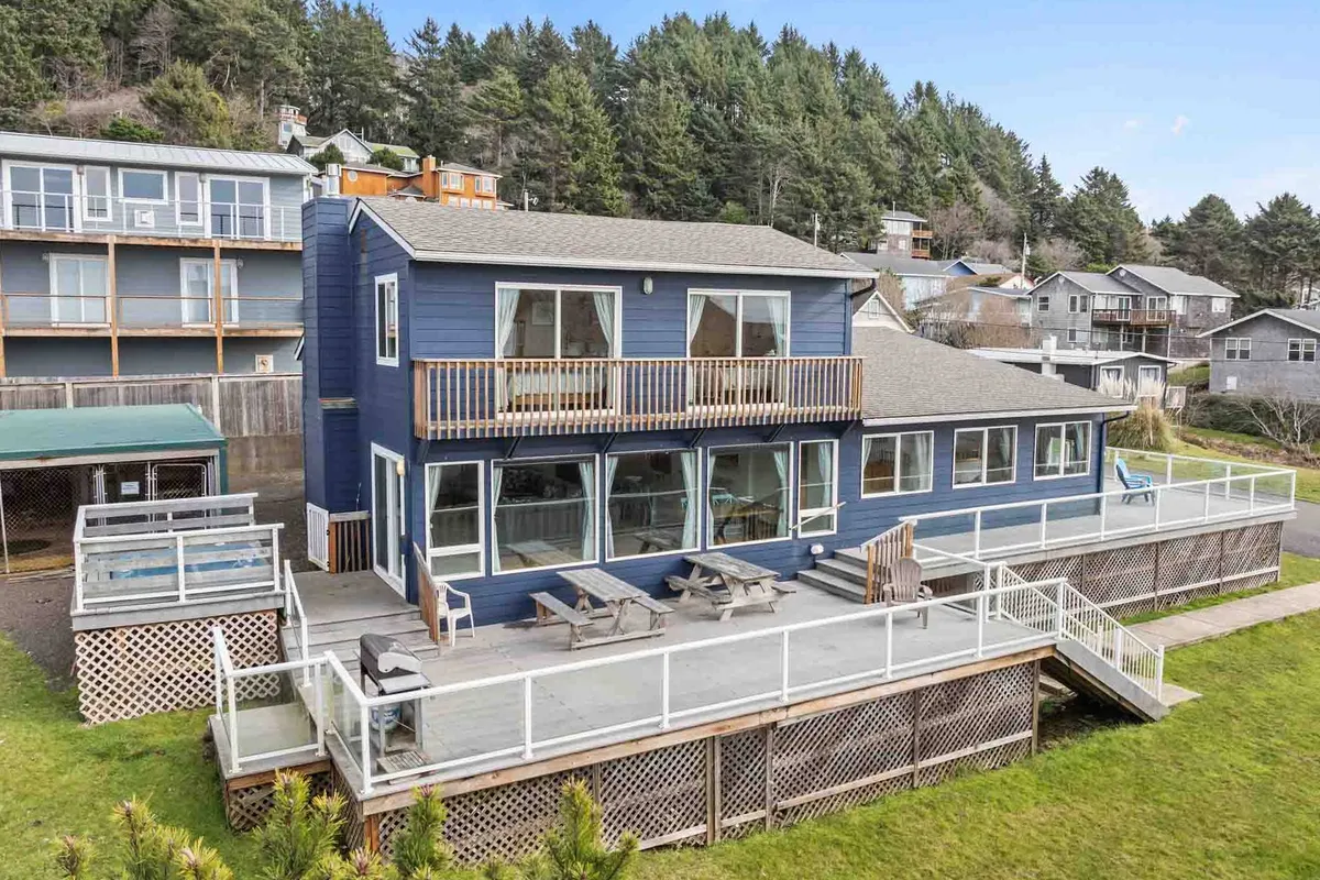 A spacious deck with expansive windows on a blue house exterior, surrounded by lush greenery. The deck is set up for outdoor dining and social gatherings and offers a direct connection to nature and the outdoors, making it ideal for group celebrations and sunset watching.