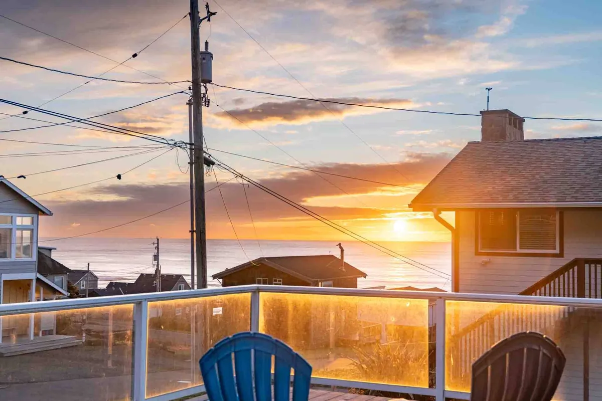 A stunning sunset view over the ocean, with a blue Adirondack chair in the foreground and the rooftops of nearby houses scattered along the coastline. The colors of the setting sun create a calm and reflective mood, inviting guests to relax and enjoy the beauty of nature after a day of coastal adventures.