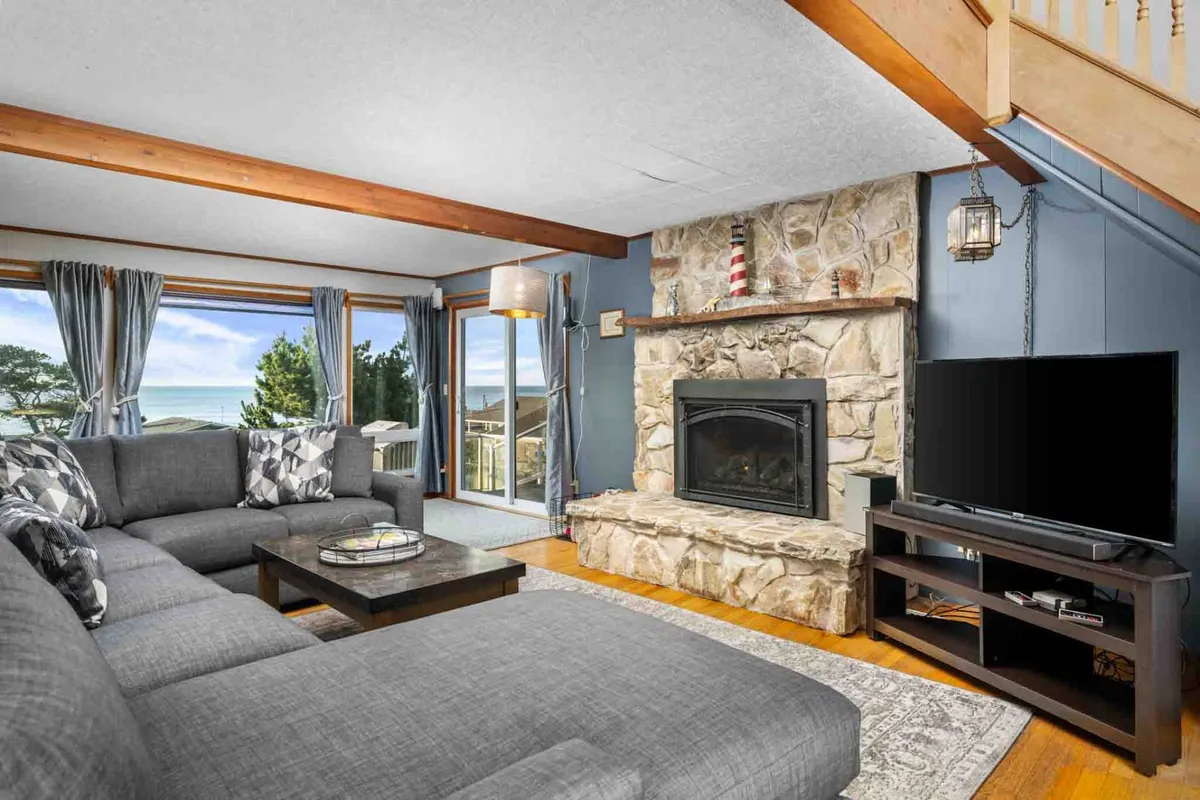 A cozy living area centered around a stone fireplace and a large sectional sofa, with wood-beamed ceilings and expansive windows looking out to the ocean. The arrangement invites family gatherings and relaxing evenings while taking in sunset views.