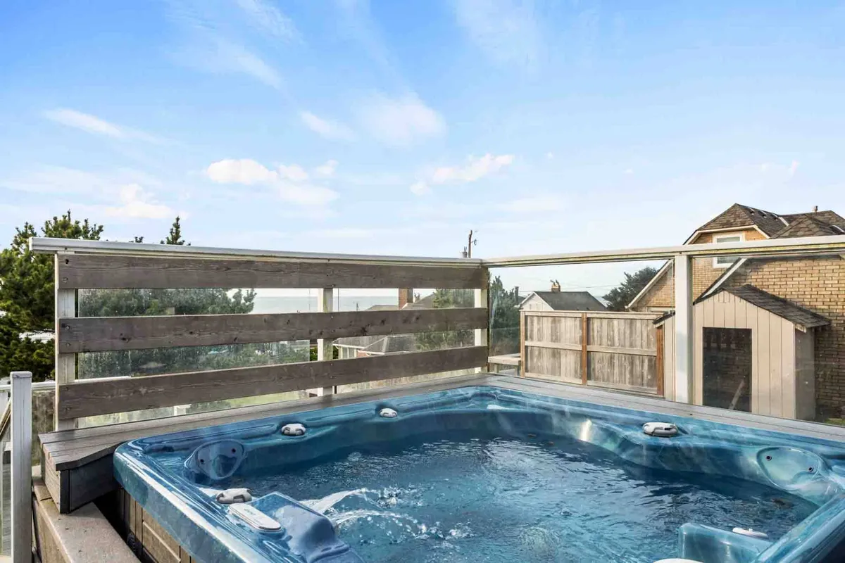 An exterior shot featuring a wind-screened hot tub on a wooden deck with views of the landscape, offering a relaxing space to unwind after beach activities. The setting is inviting, with space for group relaxation and socializing.