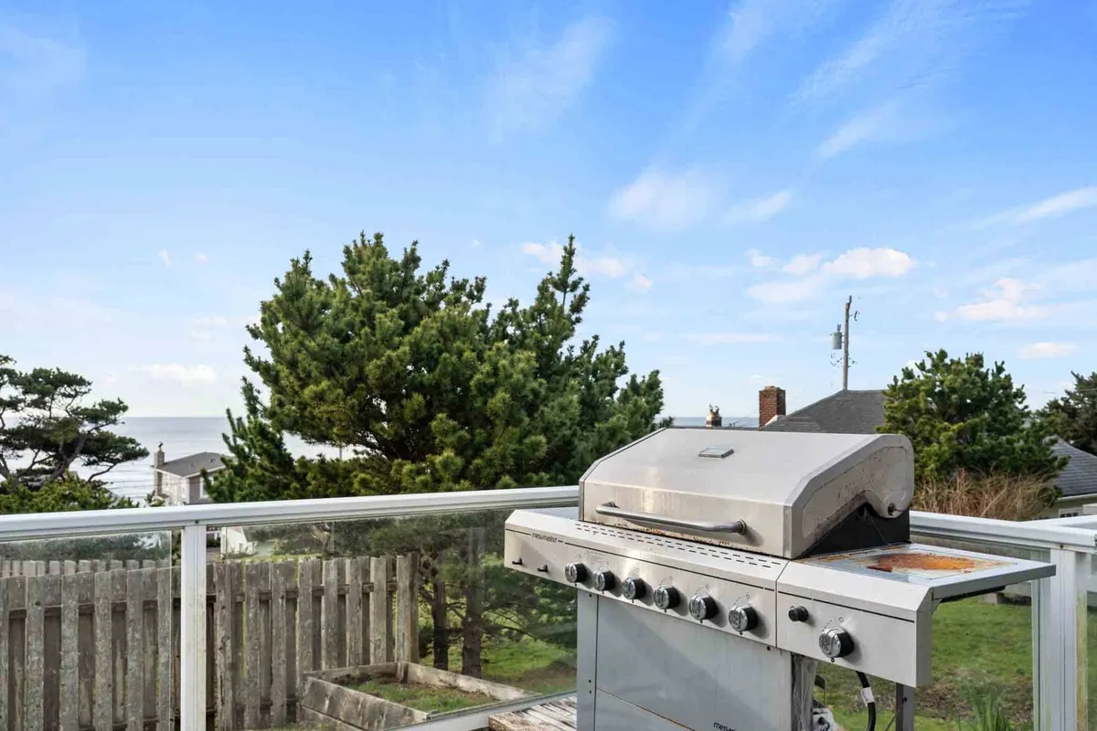 A grill set up on an outdoor patio, framed by trees and an ocean view. The scene suggests barbecuing, outdoor dining, and relaxing with friends as the sun sets, embodying the laid-back, communal spirit of a coastal lifestyle.